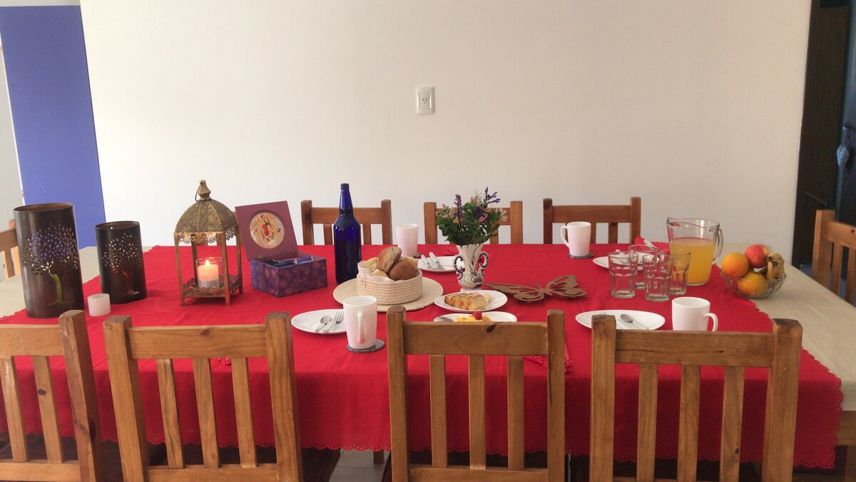 A large wooden dining table is set with a red tablecloth and is arranged to seat twelve. The table is adorned with plates of food, cups, and a fruit bowl. Decorative items include a lantern and small flowers, contributing to a welcoming dining atmosphere.