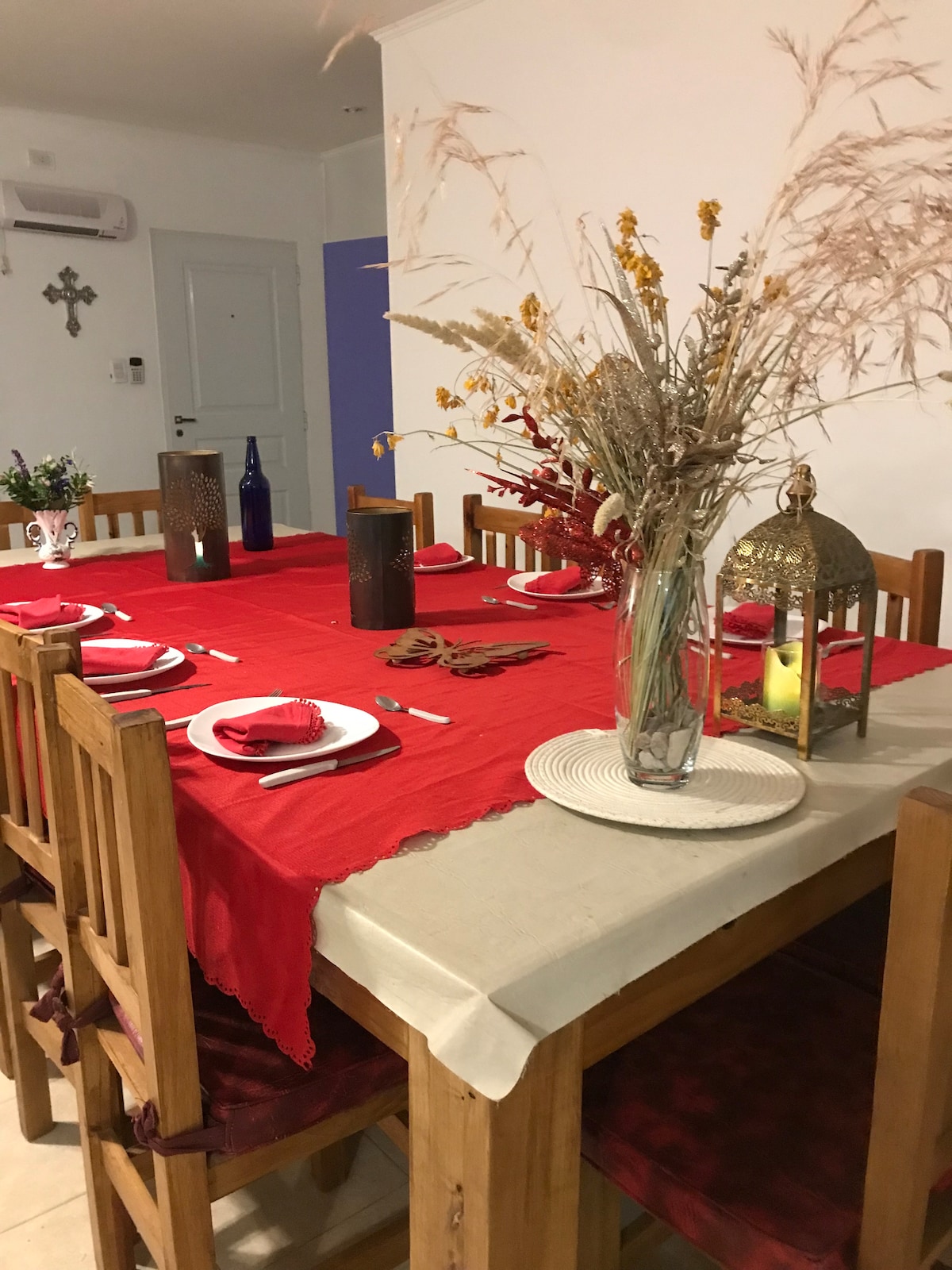 A large dining table is set for twelve, featuring a vibrant red tablecloth and neatly arranged plates and cutlery. Natural elements, including dried flowers and a decorative lantern, adorn the center of the table, adding a touch of elegance to the inviting dining space.
