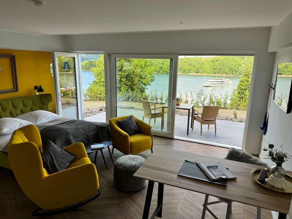 A bright living space is presented with large glass doors allowing views of the lake. Two cozy yellow swivel chairs are positioned near a table, while a spacious bed is visible in the background. A wooden table with items on it adds functionality to the area.