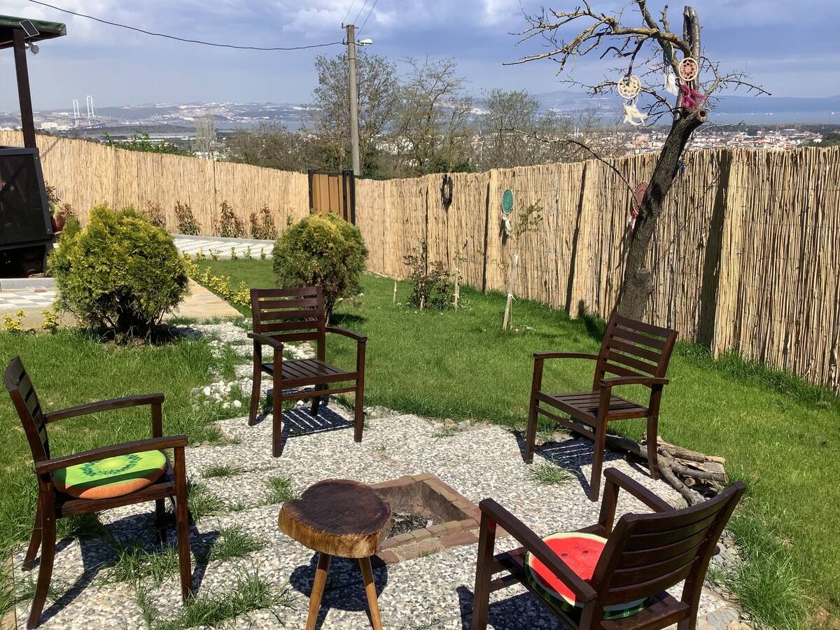 An outdoor seating area is surrounded by a neatly manicured lawn and decorative shrubs. Four wooden chairs, each featuring cushions, are arranged around a central fire pit. A rustic fence and trees provide a natural backdrop against the expansive view of the landscape.