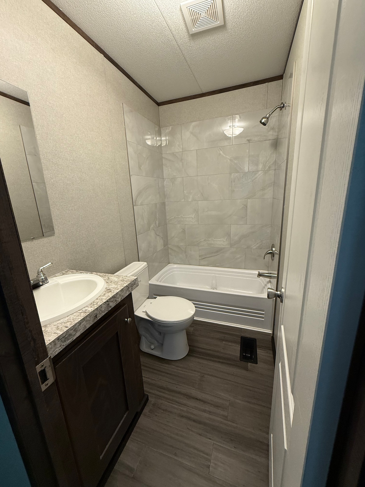 A well-appointed bathroom is highlighted by elegant marble-like tile surrounding the bathtub. A spacious sink with a granite countertop is positioned next to a large mirror. Natural light is provided by overhead lighting, enhancing the functional and tidy space.