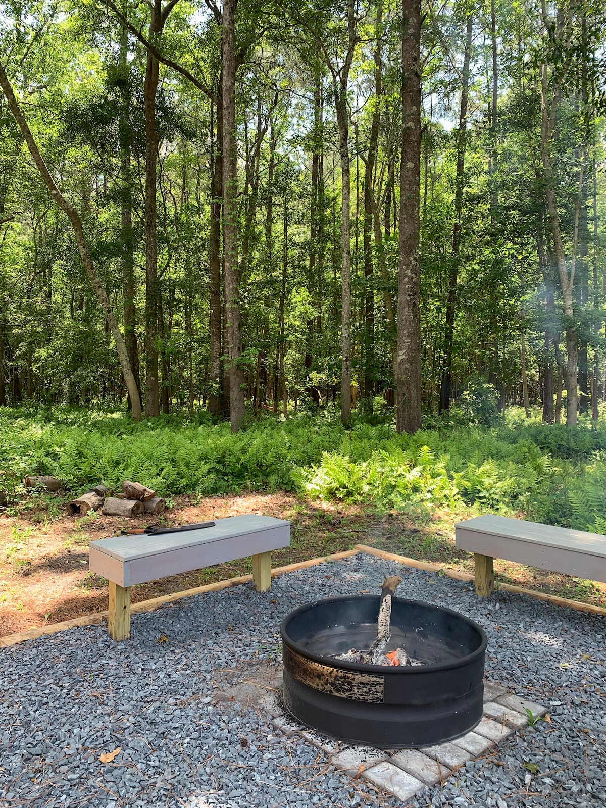 An outdoor firepit is surrounded by gravel, with two wooden benches positioned for seating. Lush green ferns and trees frame the serene area, creating a natural backdrop for relaxation. Gentle smoke rises from the firepit, indicating warmth and a cozy atmosphere.