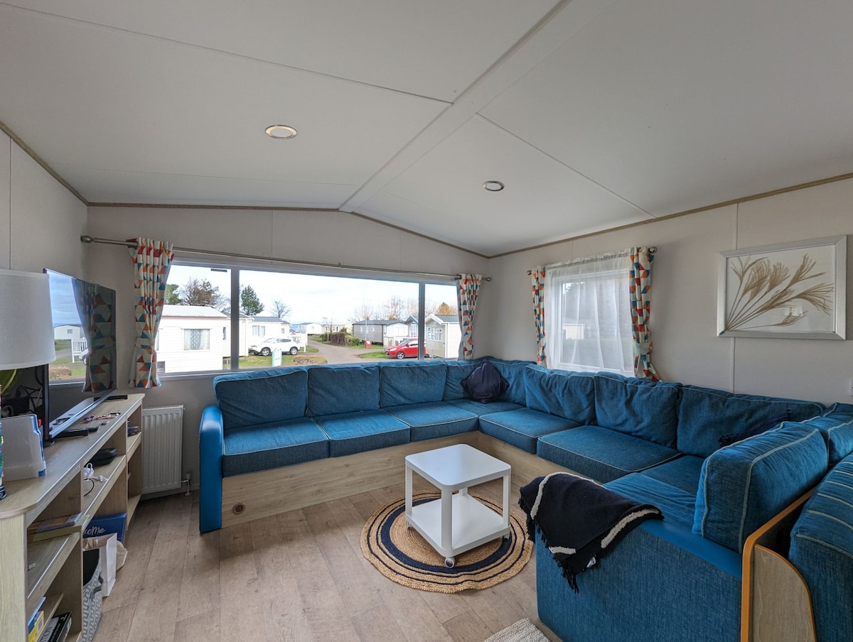 A spacious living area is highlighted by a large blue sectional sofa that accommodates several guests. A white coffee table rests in the center, and natural light filters through large windows adorned with patterned curtains. A TV stands on a console opposite the seating arrangement.