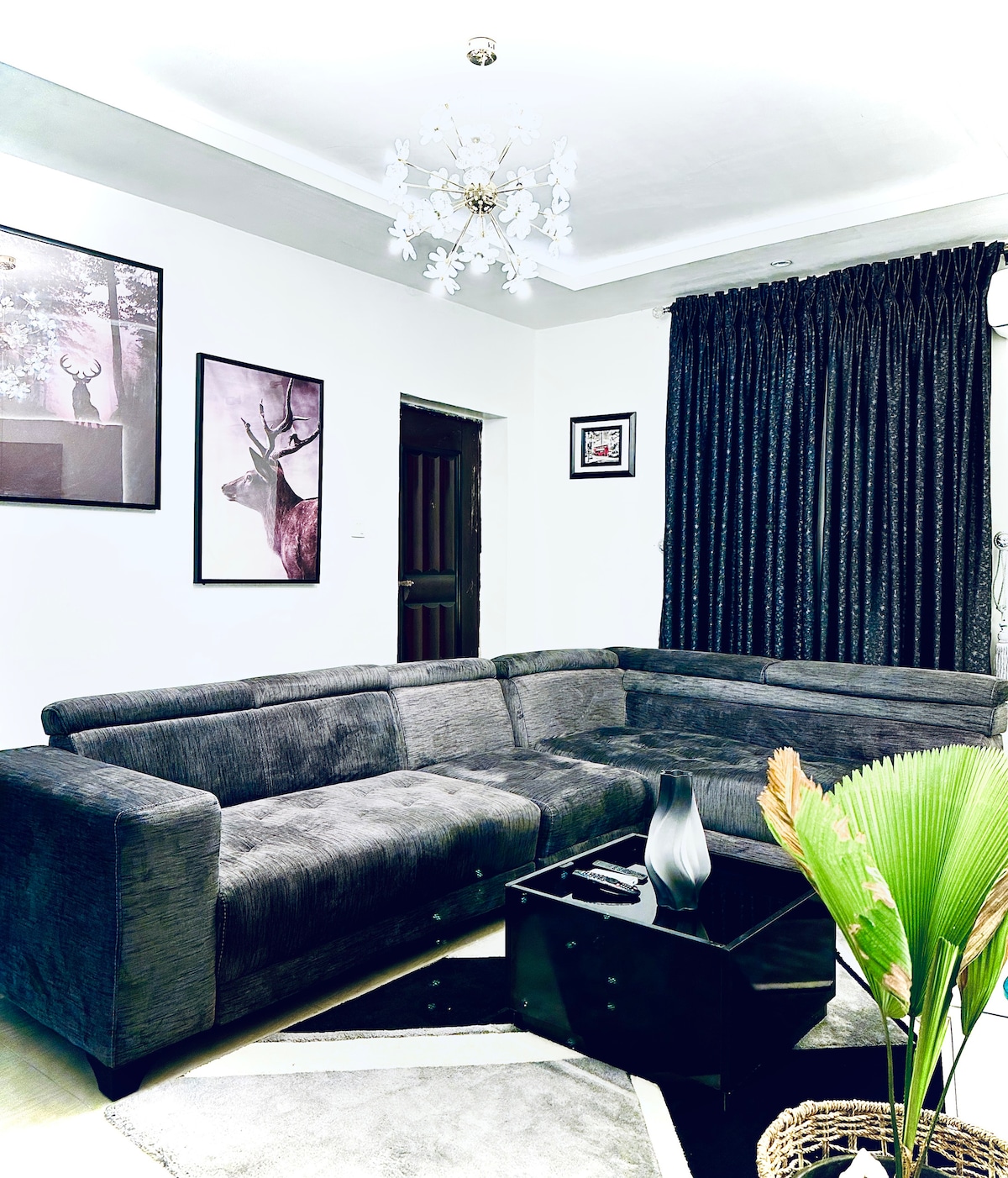 A modern living room features a plush sectional sofa arranged around a sleek coffee table. Framed art displays graceful deer against a light backdrop. Dark curtains frame the large window, and a decorative plant adds a touch of greenery to the inviting space.