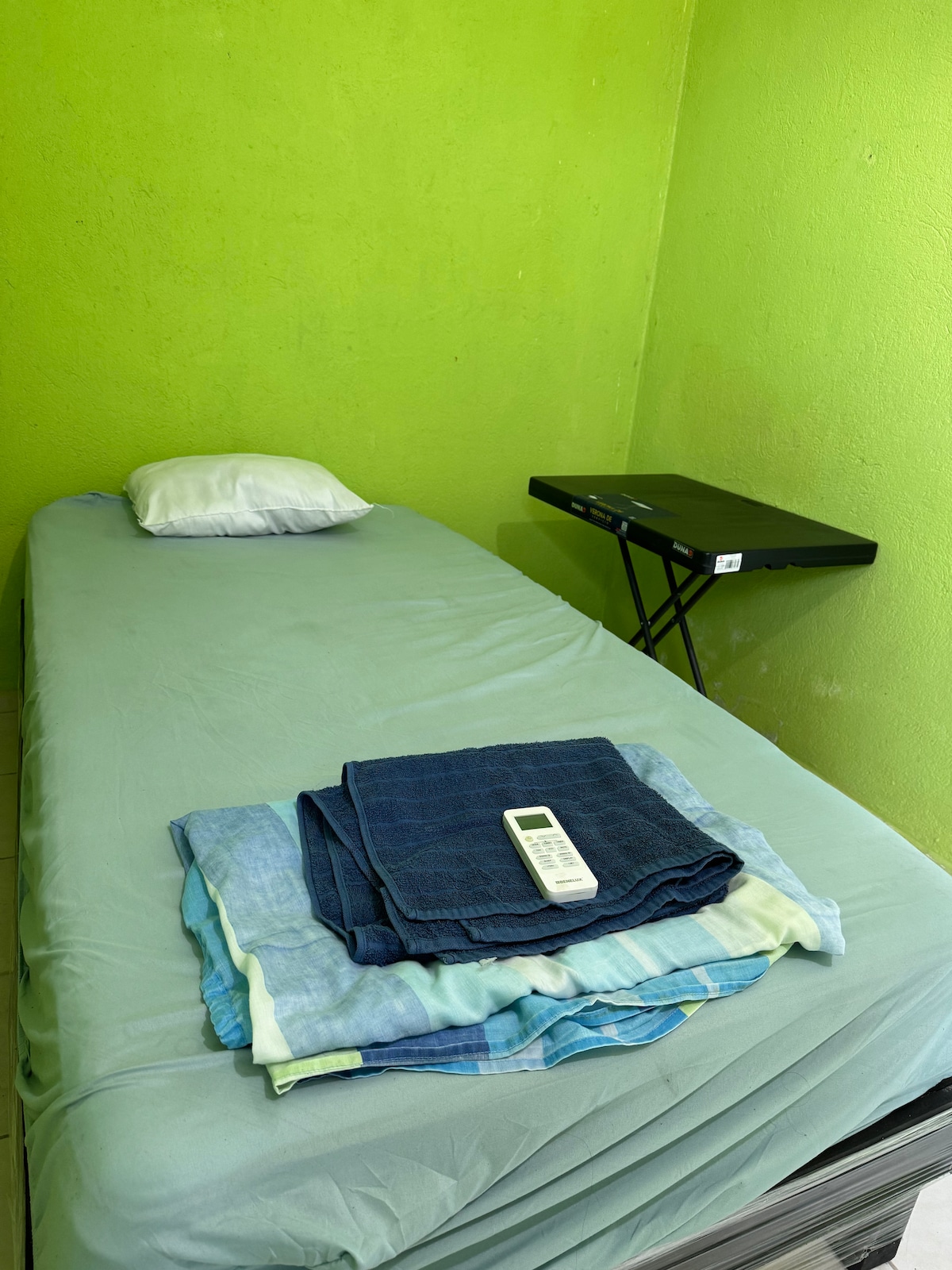 A simple sleeping area features a narrow bed with a light green sheet and a white pillow. A stack of neatly folded towels sits on the bed. In the corner, a small table offers additional space.