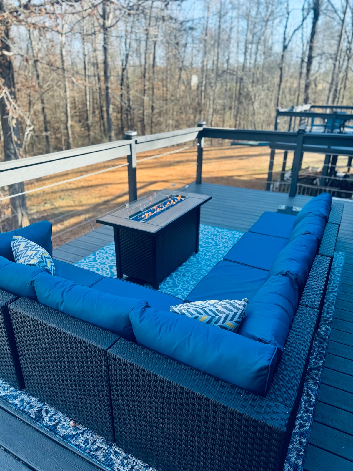 A deck area features a large sectional seating arrangement with blue cushions, positioned around a central fire table. The outdoor rug adds texture, while the surrounding tree line provides a serene backdrop.