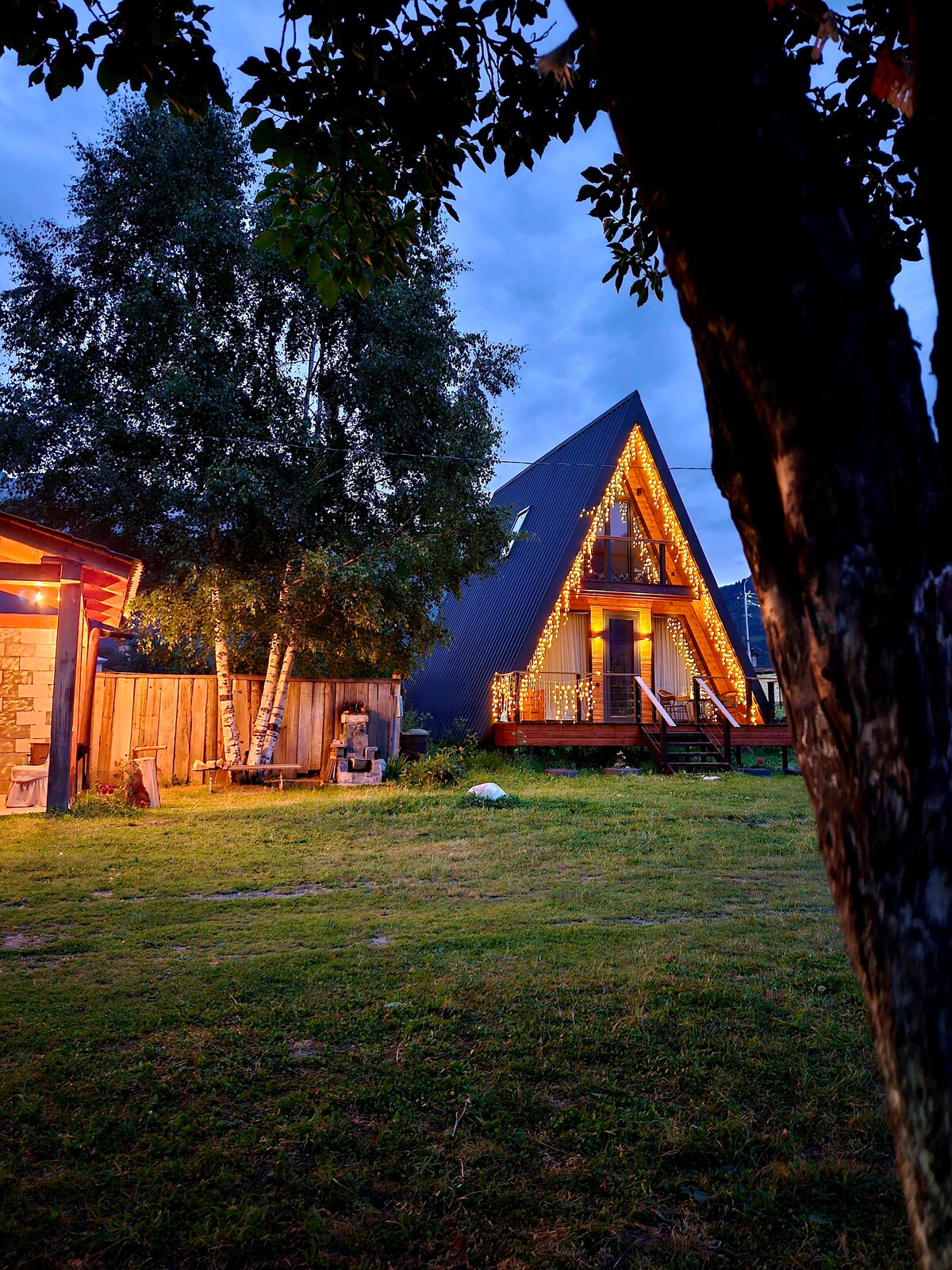 A charming A-frame house is set in a grassy area, illuminated by outdoor lighting. The triangular roof is complemented by decorative string lights, creating a welcoming ambiance. Nearby, a wooden patio invites relaxation amid trees, enhancing the home's rustic character.