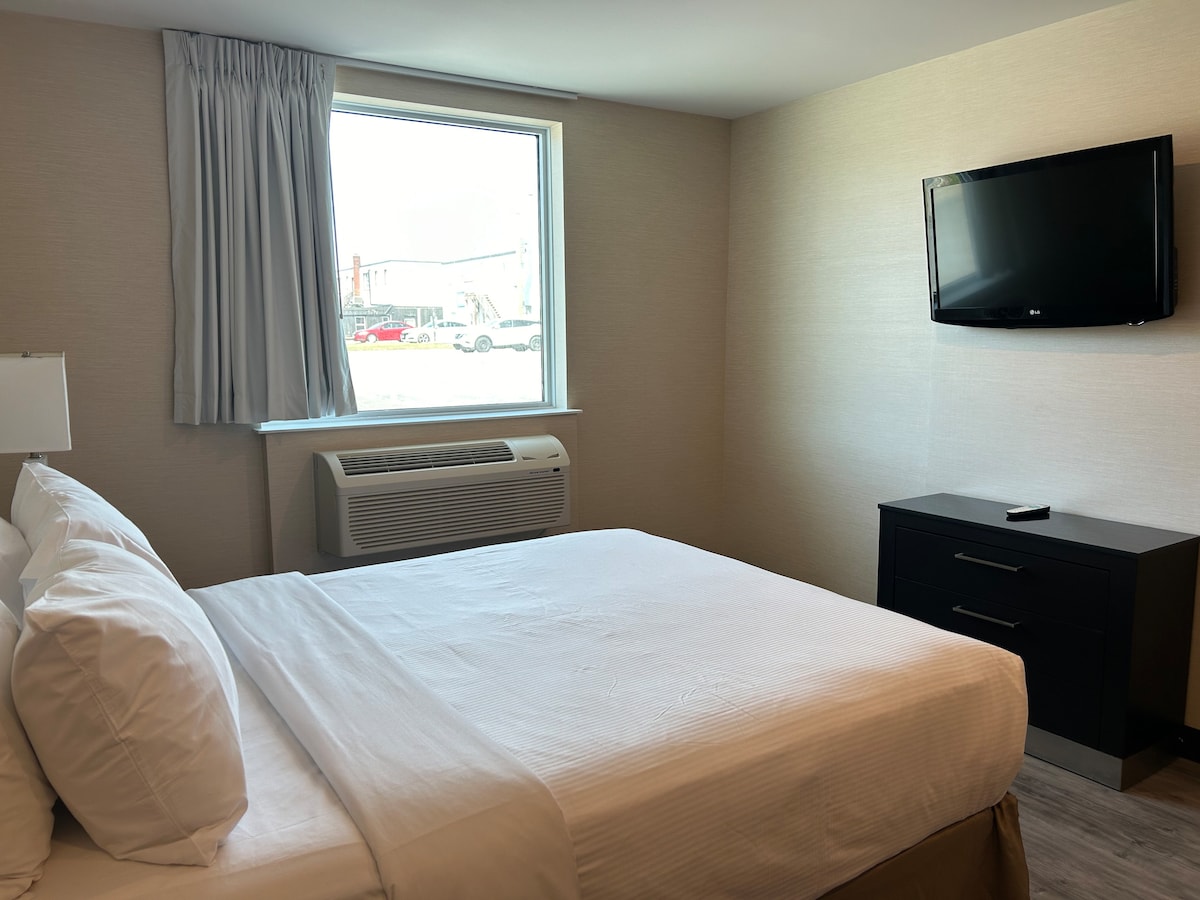 A well-lit sleeping area features a spacious bed with crisp white linens. A window is positioned nearby, allowing natural light to fill the room. An air conditioning unit can be seen beneath the window, while a black dresser holds a flat-screen TV mounted on the wall.