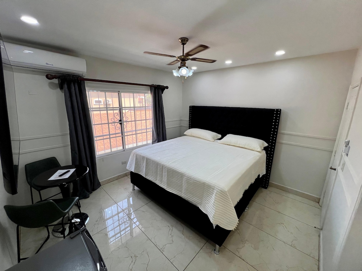 A spacious bedroom features a black upholstered bed with white bedding. Two windows allow natural light to fill the room, complemented by ceiling lights and a ceiling fan. A small table and two chairs are positioned nearby, creating a cozy seating area.