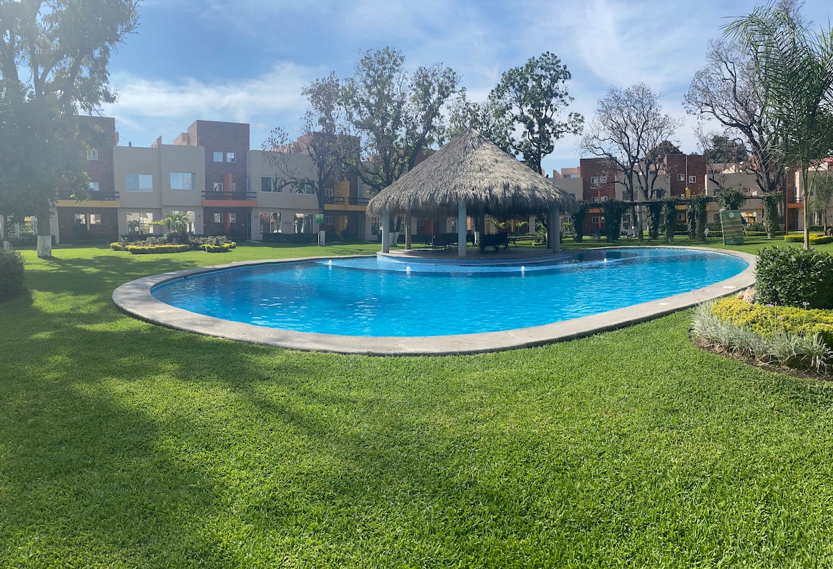 A large swimming pool is surrounded by lush greenery and manicured lawns, offering an inviting turquoise color. A thatched-roof palapa provides shaded seating near the water, while spacious residential buildings are visible in the background.