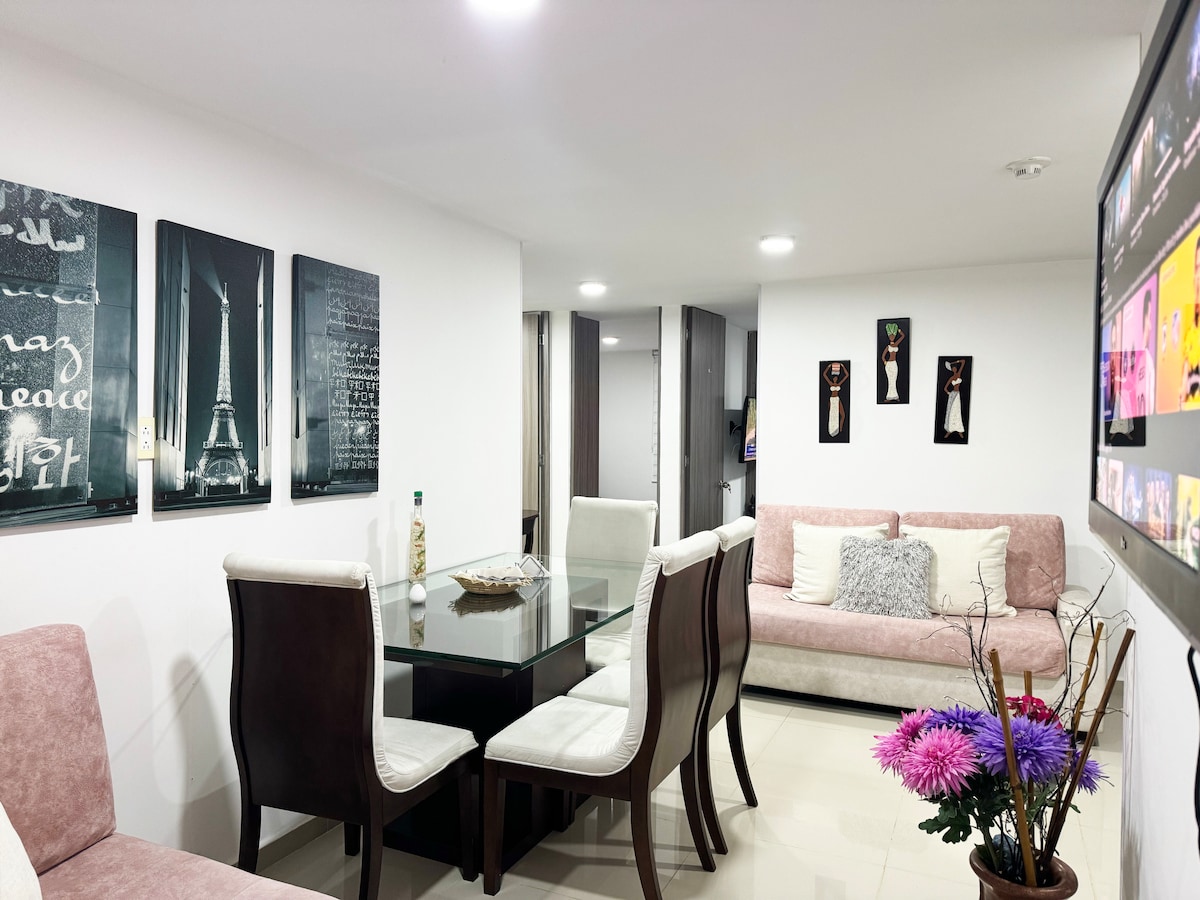 A modern living area features a glass dining table surrounded by upholstered chairs. Soft pink and white furnishings complement the decor. Wall art depicting iconic landmarks adds character. A flat-screen TV is mounted, and a decorative vase of flowers provides a touch of color.