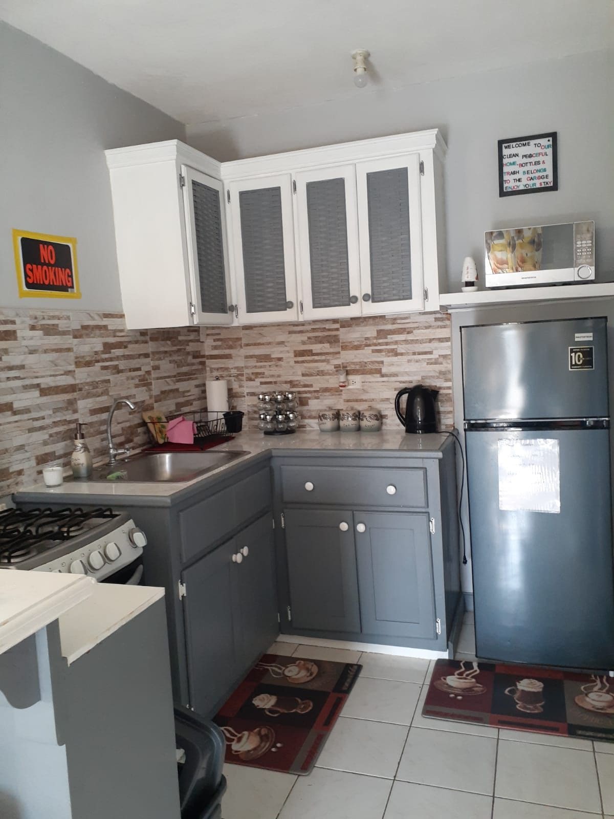 A compact kitchen features gray cabinetry and a tiled backsplash. Essential appliances, including a stove, microwave, and refrigerator, are present. Counter space is utilized for meal preparation, complemented by a metal kettle and dishware. Flooring is tiled, with a patterned rug adding a decorative touch.