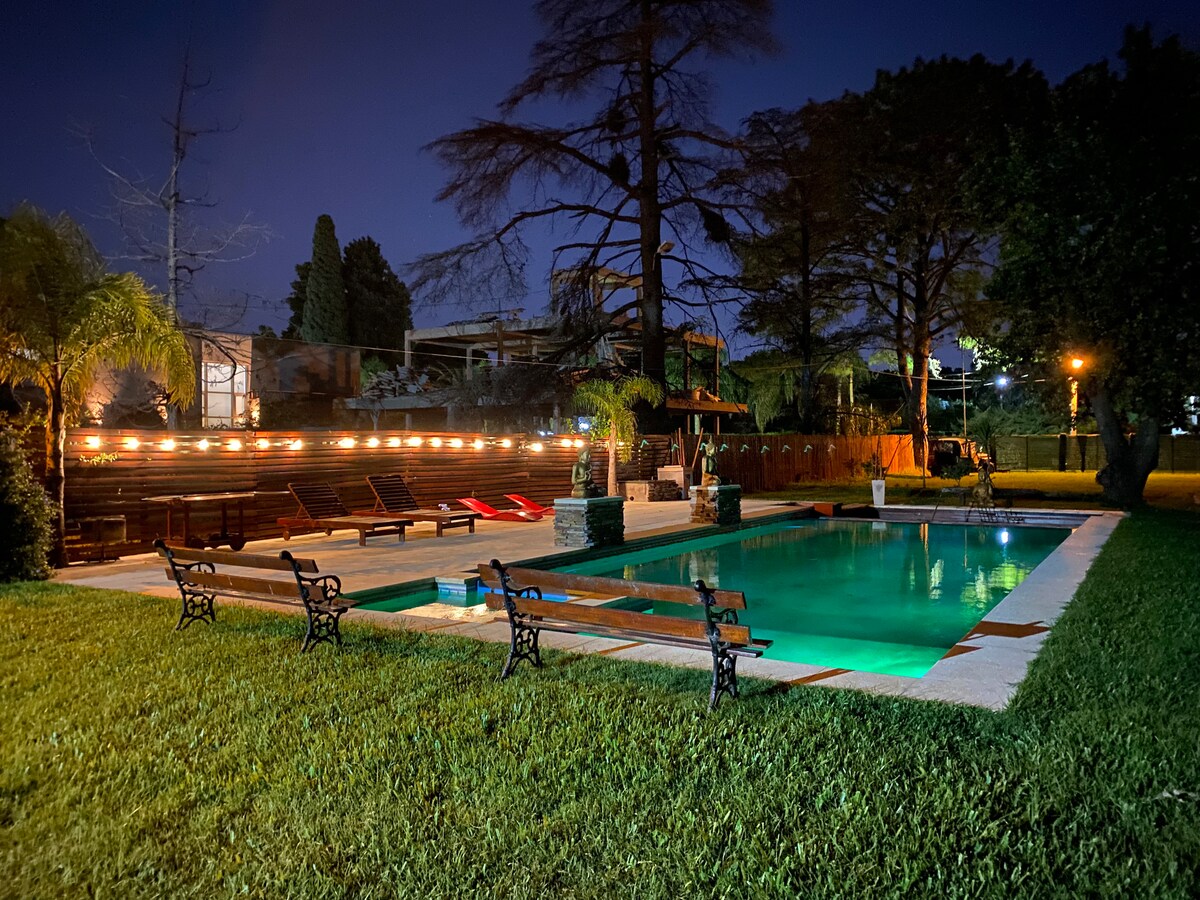 A well-lit swimming pool is surrounded by landscaped greenery, with lounge chairs positioned nearby. Soft lighting enhances the inviting atmosphere of the outdoor space, creating a peaceful setting for relaxation. The pool water reflects the serene surroundings under the night sky.