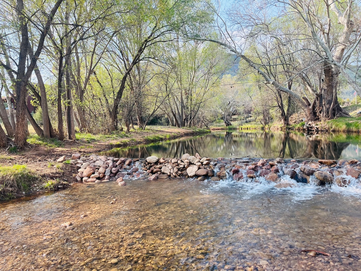 A serene scene of the East Verde River features gentle water flow over smooth stones, bordered by lush greenery and trees. Reflections of the surrounding nature can be seen on the water's surface, enhancing the peaceful ambiance of this natural setting.