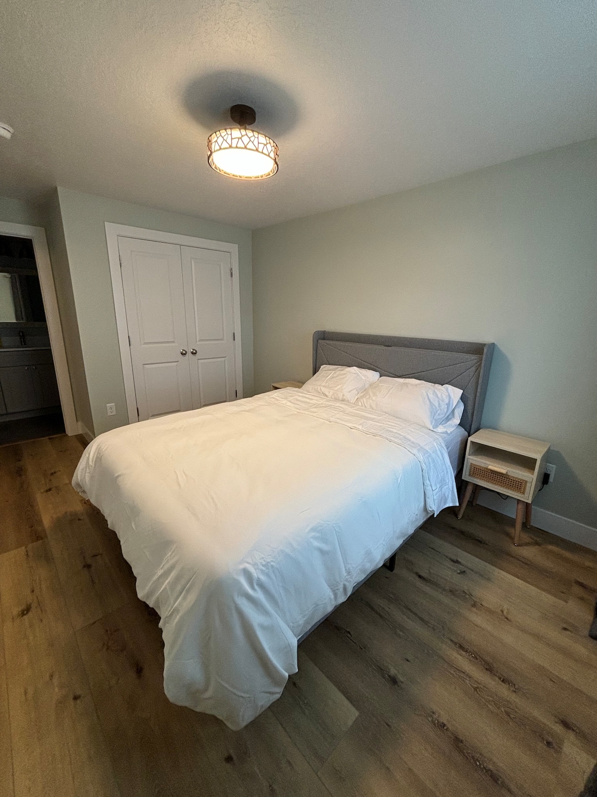 The bedroom features a queen size bed dressed in white linens, flanked by two nightstands. A light fixture hangs from the ceiling, and natural light enters through a nearby closet door, providing a soft ambiance. The room is finished with light green walls and wood flooring.