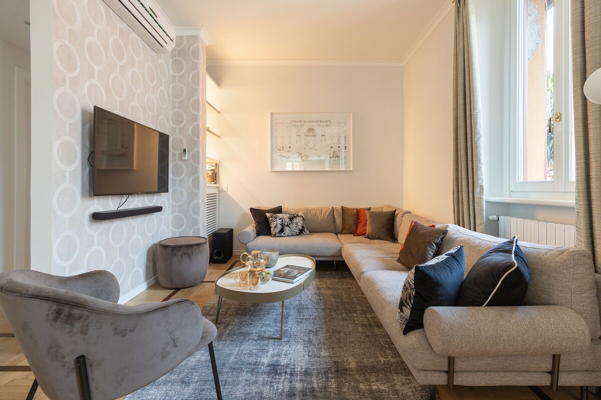 A modern living area is presented with a spacious sectional sofa adorned with decorative pillows. A sleek coffee table is positioned in front, accompanied by a soft area rug. Natural light enters through a nearby window, highlighting the contemporary decor and wall-mounted TV.