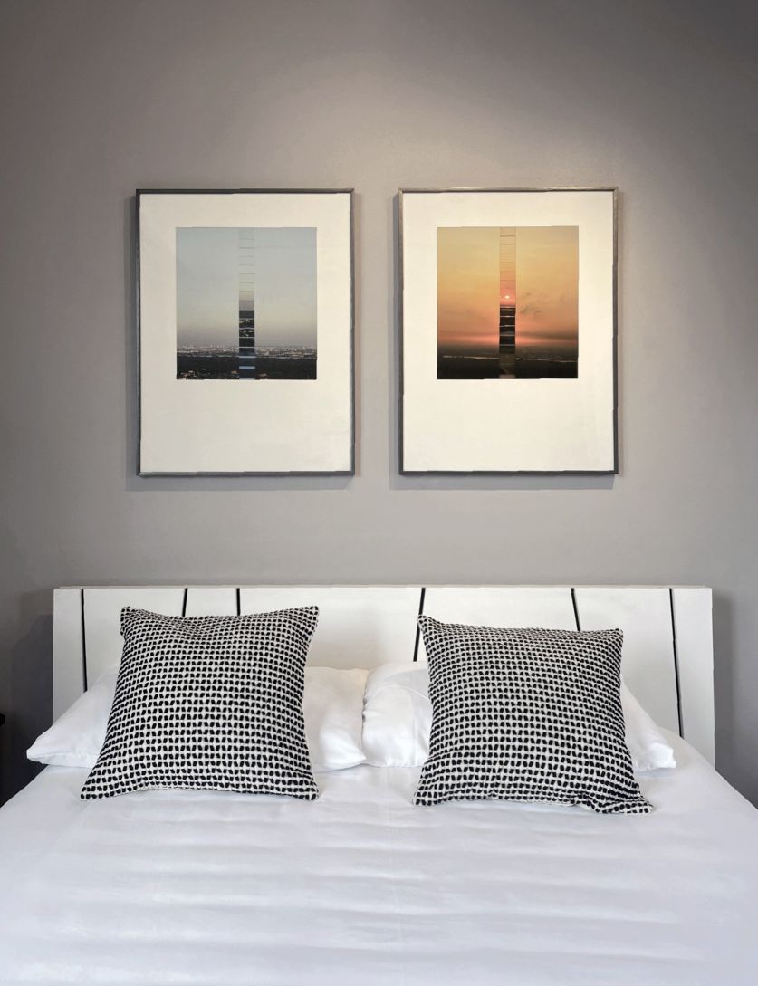 A spacious bed is centered against a muted gray wall, adorned with two framed artworks showcasing contrasting scenic views. Decorative pillows with a patterned design are positioned neatly on the bed. Natural light illuminates the room, creating a serene atmosphere.