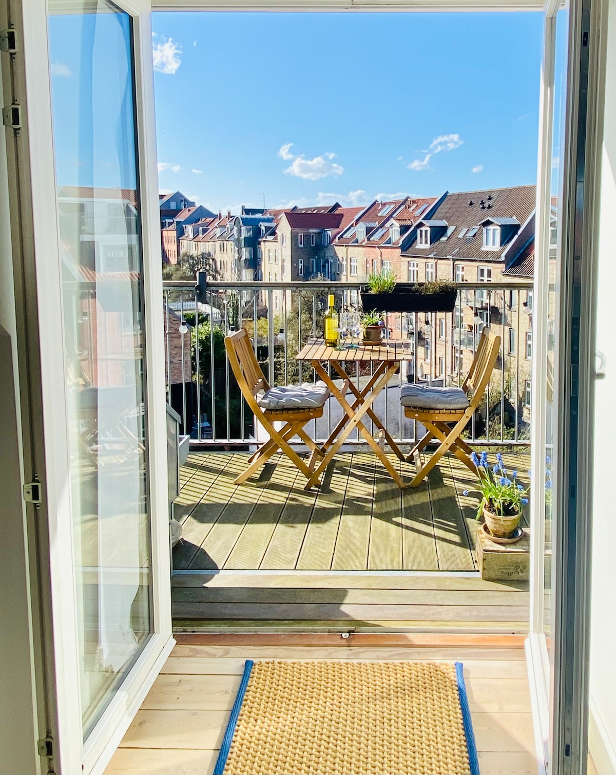 A private balcony is visible, featuring a small round table with two wooden chairs. Potted plants are placed nearby, and the view showcases neighboring buildings under a clear blue sky with scattered clouds.