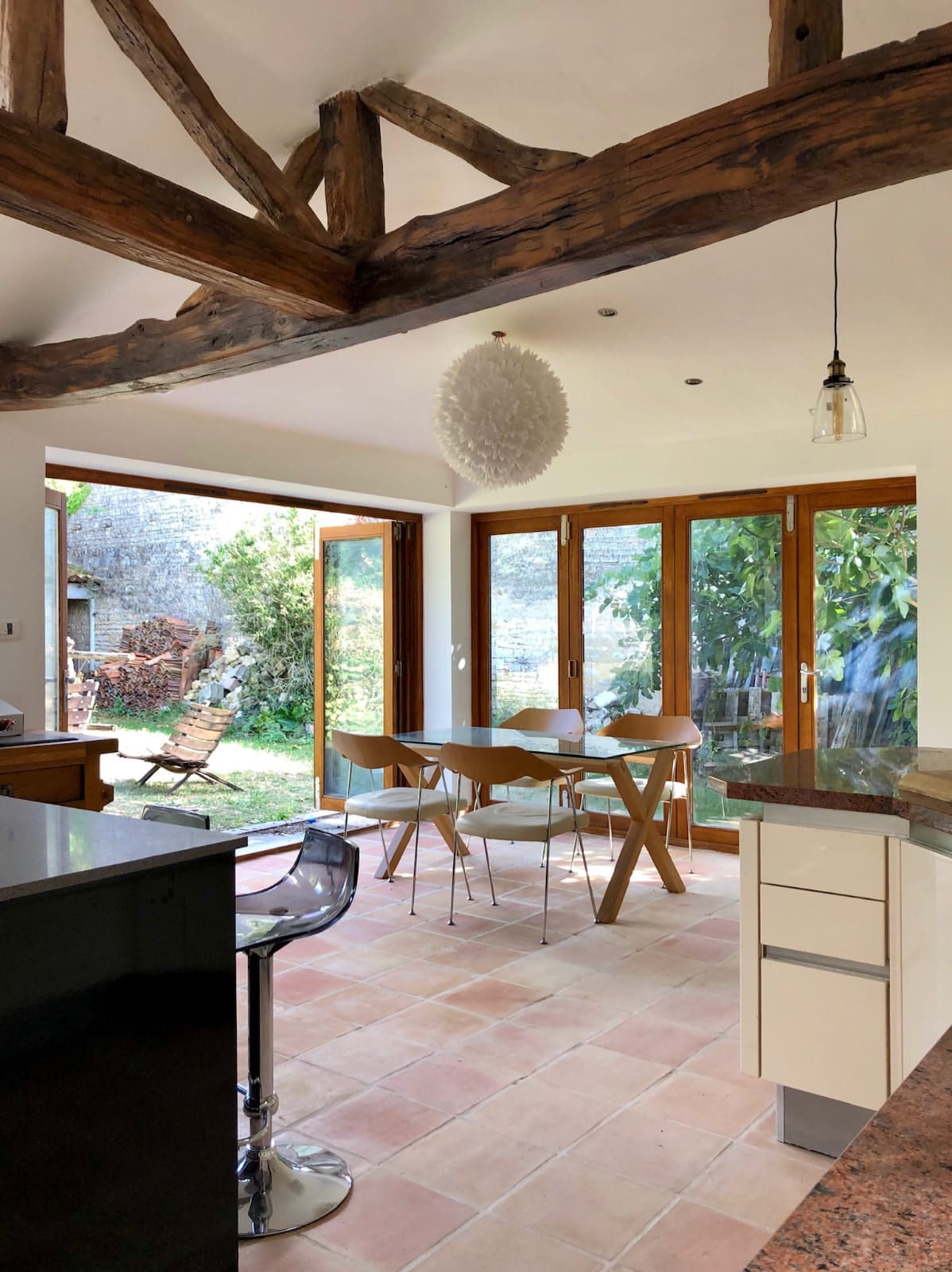 An open-plan kitchen and dining area features wooden beams and a modern pendant light. Glass doors lead to a garden, allowing natural light to fill the space. A wooden dining table is surrounded by four chairs, complemented by sleek bar stools at the kitchen counter.