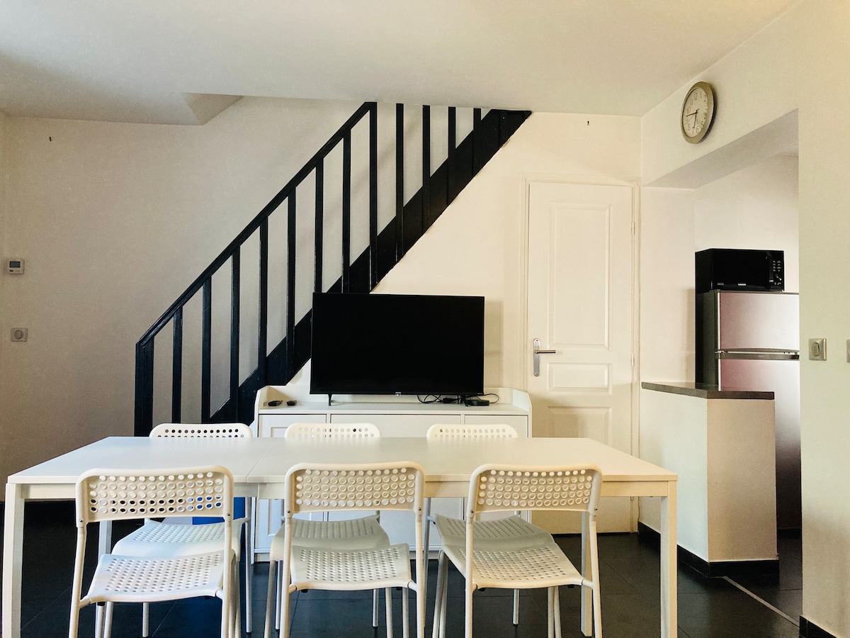 The living area features a modern dining table with four white chairs, positioned across from a flat-screen television. A staircase leads to the upper level, while a compact kitchen area is visible to the side, complemented by stainless steel appliances.