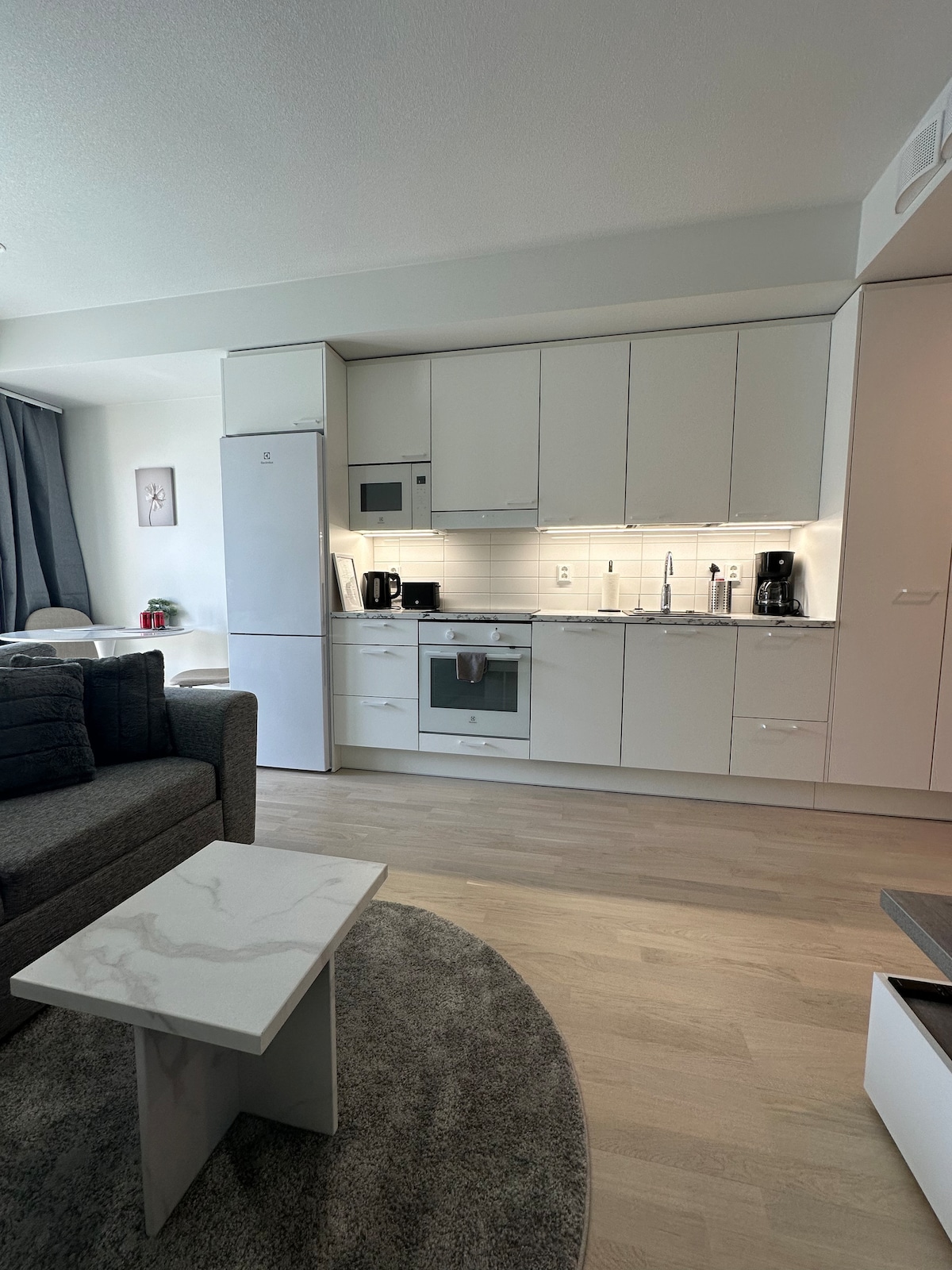 A modern kitchenette is displayed with sleek white cabinets and a light countertop. Kitchen appliances, including a refrigerator and microwave, are conveniently arranged. A cozy seating area, with a dark sofa, is visible in the background, along with a soft area rug under a small coffee table.