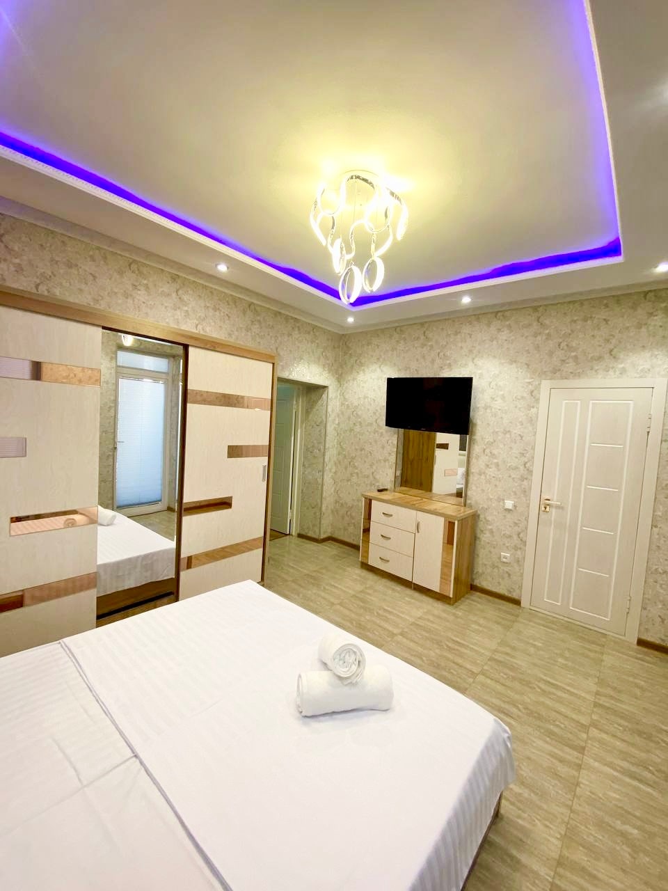 The room features a spacious bed with crisp white linens, complemented by a modern chandelier with soft lighting. A flat-screen TV is mounted on the wall, while a wooden dresser offers storage. Neutral wall decor and large mirrored closets enhance the sense of space.