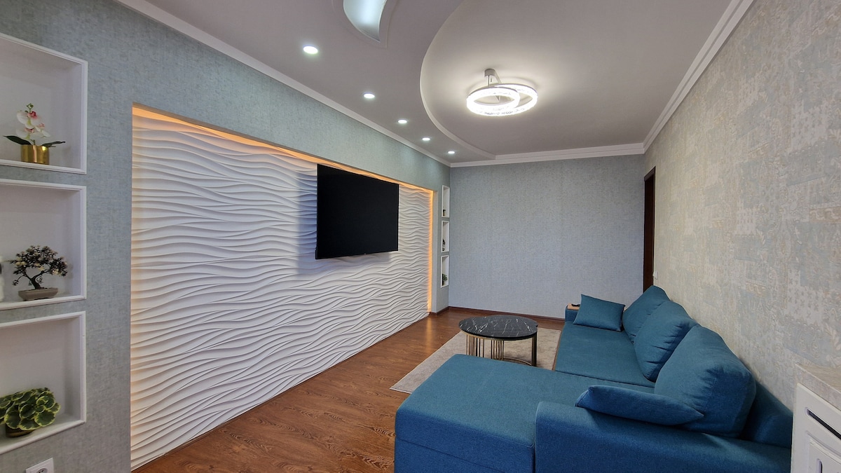 A comfortable living area is highlighted by a textured feature wall with a mounted television. A modern coffee table is positioned in front of a spacious blue sofa, with soft lighting provided by recessed ceiling fixtures. A warm wood floor complements the inviting space.
