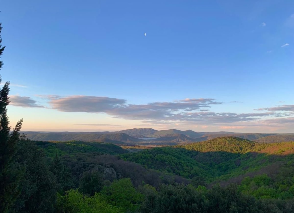 A panoramic view of rolling green hills and valleys stretches under a clear sky. A subtle glow from the setting sun casts soft shadows across the landscape, while a small moon is visible in the blue sky.