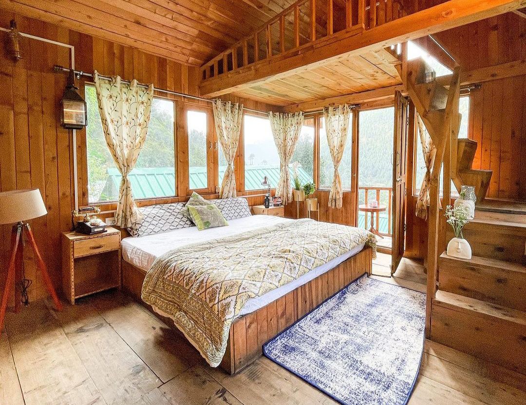 A wooden bedroom features a spacious bed adorned with a patterned blanket. Sunlight streams through multiple windows, creating a bright ambiance. A staircase leads to a loft area, while a cozy rug adds warmth to the wooden flooring. Natural wood elements dominate the interior design.