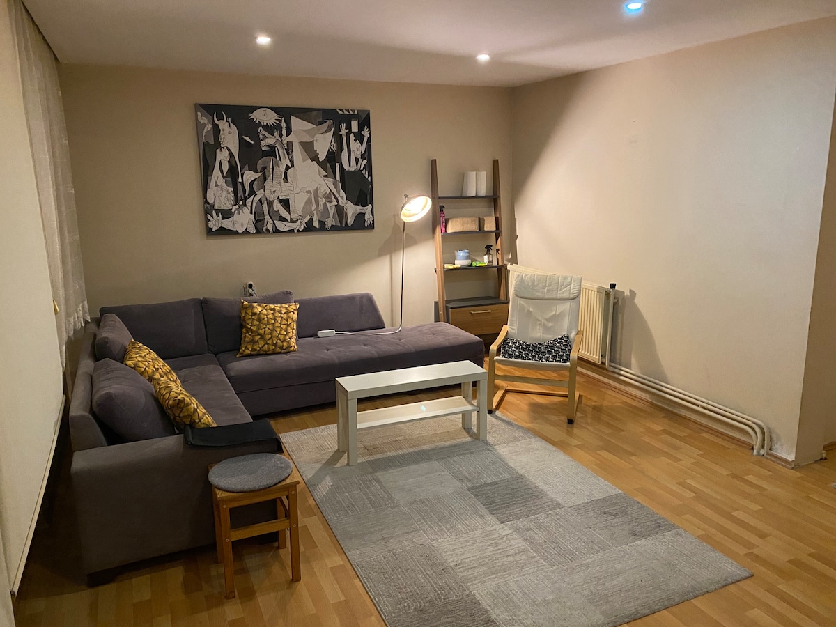 A comfortable living area is furnished with a large sectional sofa featuring decorative cushions. A light stands next to a small table, while a wooden chair and a stool provide additional seating. The space is illuminated by soft overhead lights and has a warm wooden floor.