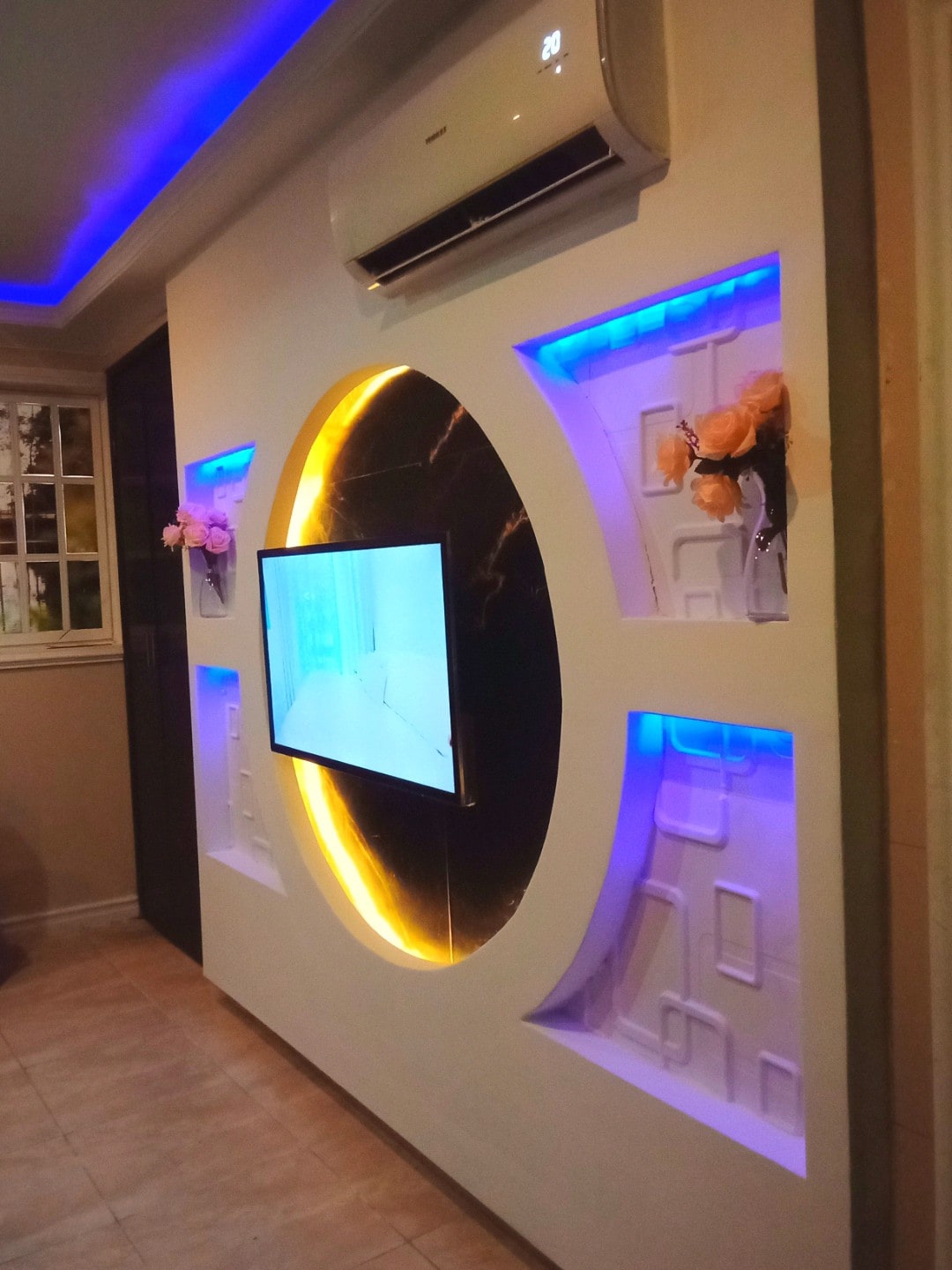 A modern living area features a sleek wall design with a flat-screen TV centered within a circular frame. Soft blue and warm yellow lighting accentuates the wall's texture, complemented by decorative elements at the edges. An air conditioning unit is mounted above for comfort.