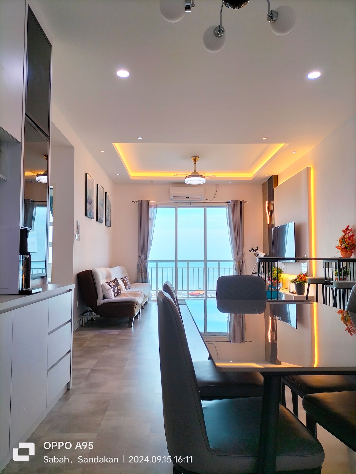 A modern living space showcases a dining area with a sleek table and chairs, adjacent to a cozy seating area featuring a sofa. Large glass doors lead to a balcony, offering a view of the surrounding area. Soft lighting enhances the contemporary design.