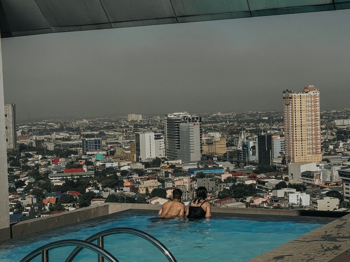 An infinity pool is observed at the rooftop level, offering panoramic views of the city skyline. Two individuals are seen relaxing in the water, overlooking a mix of urban buildings and green spaces. The horizon features a hazy skyline, suggesting a warm afternoon.