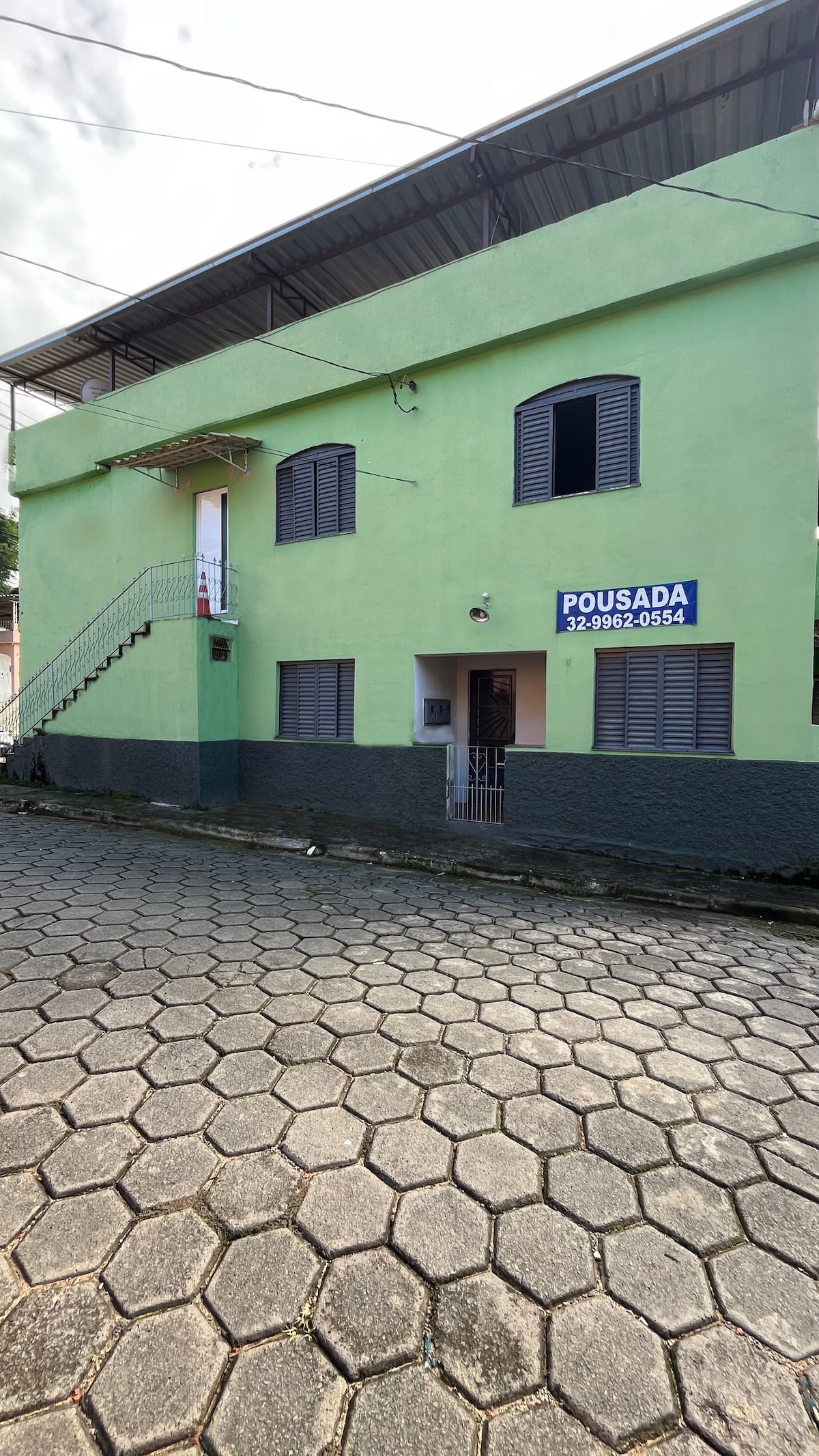 A green, two-story building can be seen, featuring multiple windows with gray shutters. A metal staircase leads to the upper level on one side. The entrance is marked by a sign displaying the property name and contact information. The surrounding area includes paved stone pathways.