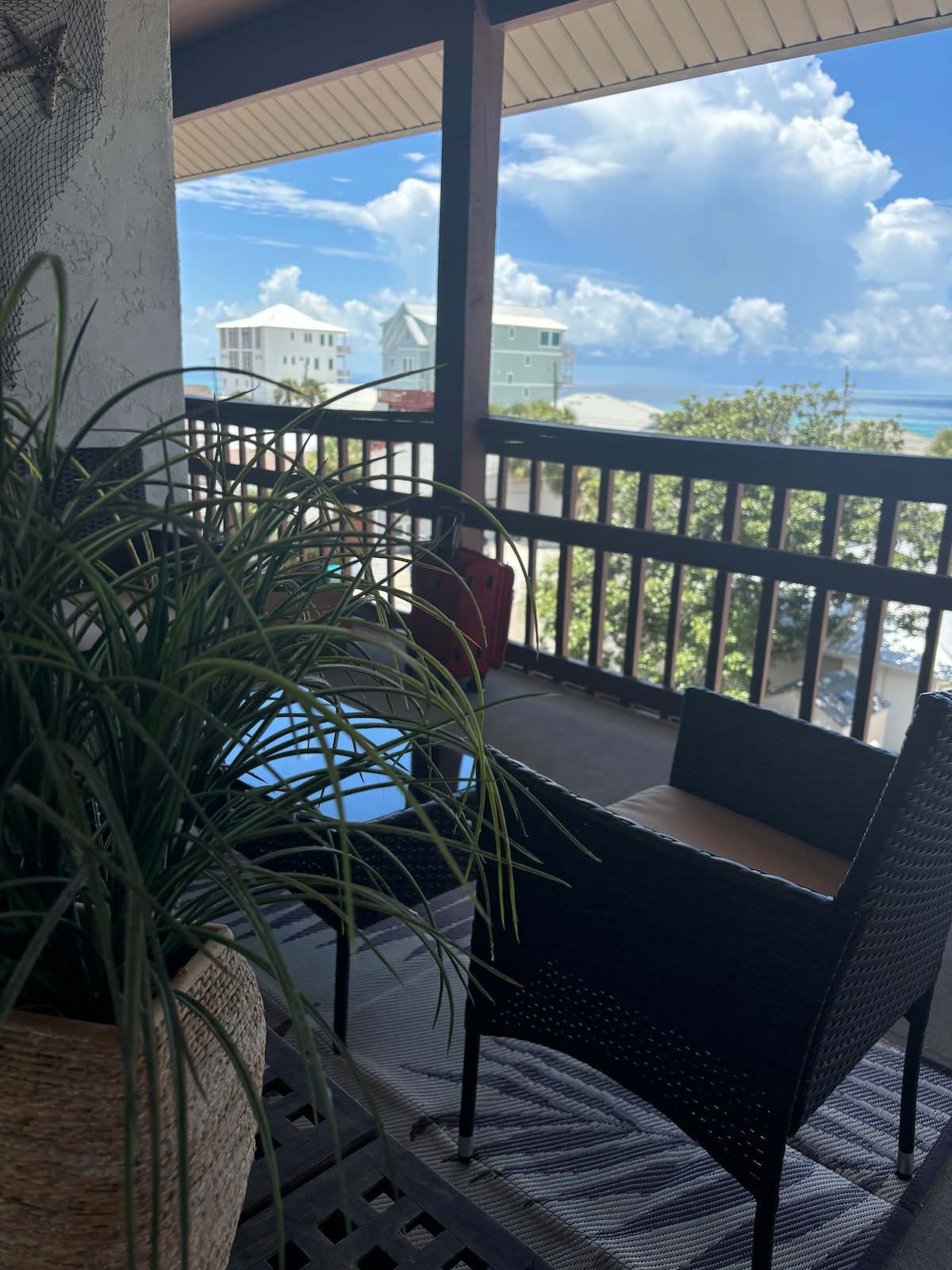 A balcony area featuring a small round table and a wicker chair. A potted plant adds a touch of greenery. The view of the ocean and clouds is visible through a railing, hinting at a coastal atmosphere.