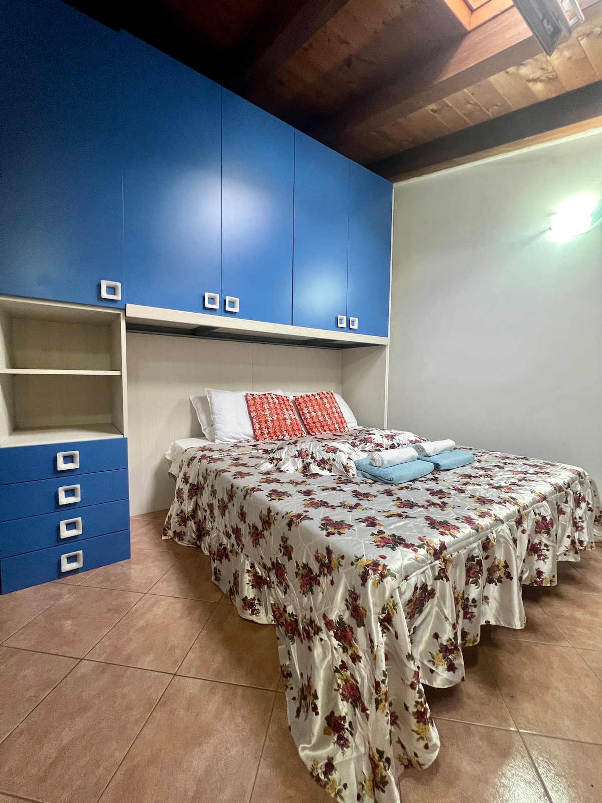 A cozy sleeping area is presented with a spacious bed adorned with a floral bedspread. Two vibrant cushions are placed at the head of the bed, while built-in blue cabinets and a small dresser provide storage. Light reflects softly from the ceiling, contributing to the room's comfort.