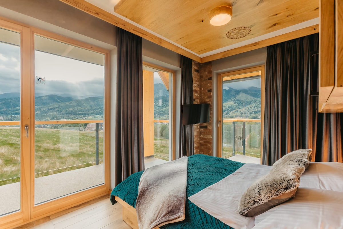 A spacious bedroom features floor-to-ceiling windows that provide views of the surrounding mountains. A large bed with decorative pillows is complemented by dark curtains. Natural light brightens the room, and a flat-screen television is mounted on the wall.