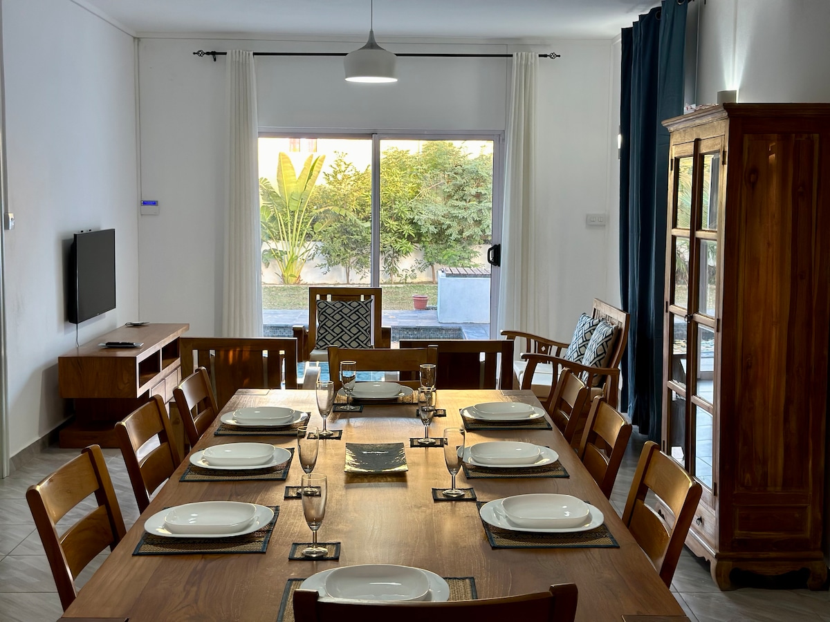 A large dining table is set with plates and glassware, surrounded by wooden chairs. Natural light floods the room through a large window, showcasing a view of the garden. A sideboard and television are positioned in the background, enhancing the open layout.