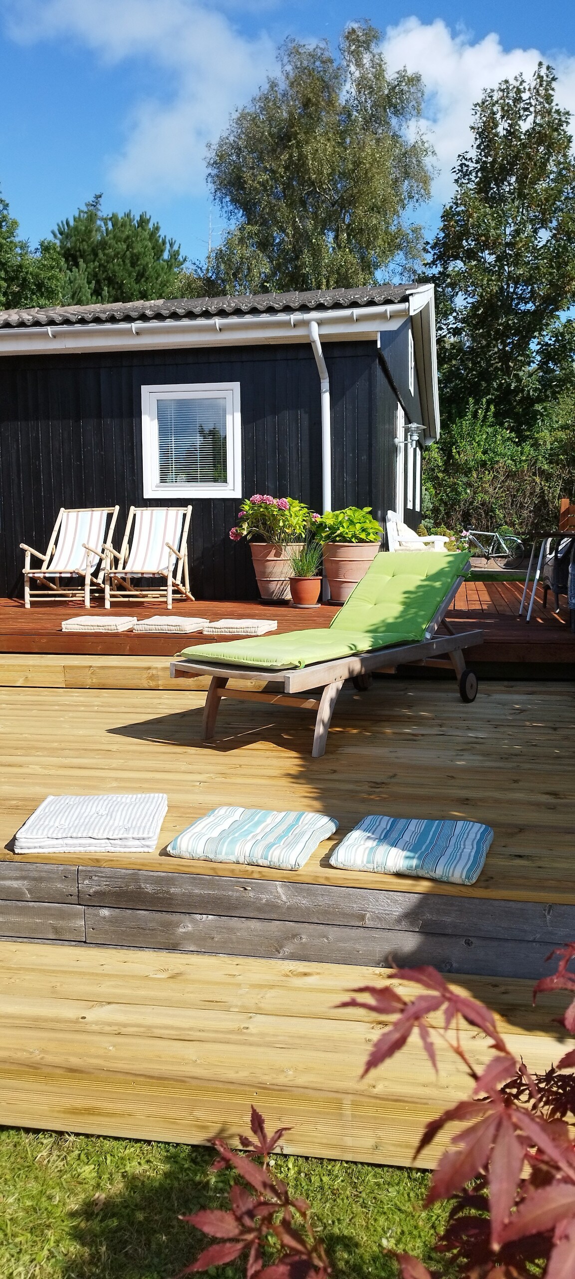 A sun-drenched terrace features a green lounge chair and two striped deck chairs. Potted plants enhance the outdoor ambiance, with a wooden deck extending from the house's exterior. A glimpse of the surrounding greenery can be seen in the background.
