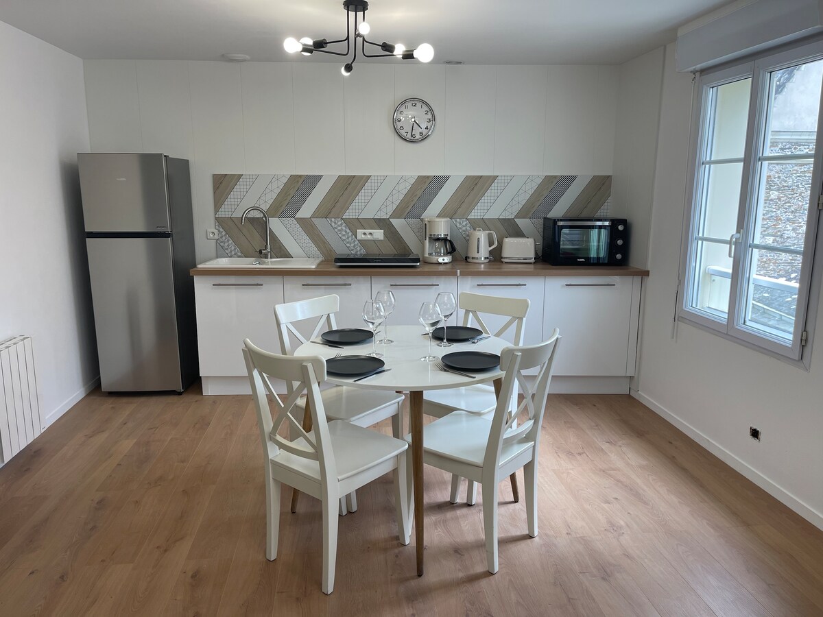 A modern kitchen is visible, featuring a round dining table set for four with white chairs. Essential appliances, including a refrigerator and microwave, are neatly arranged along the wall, complemented by a stylish backsplash. Natural light enters through a window.