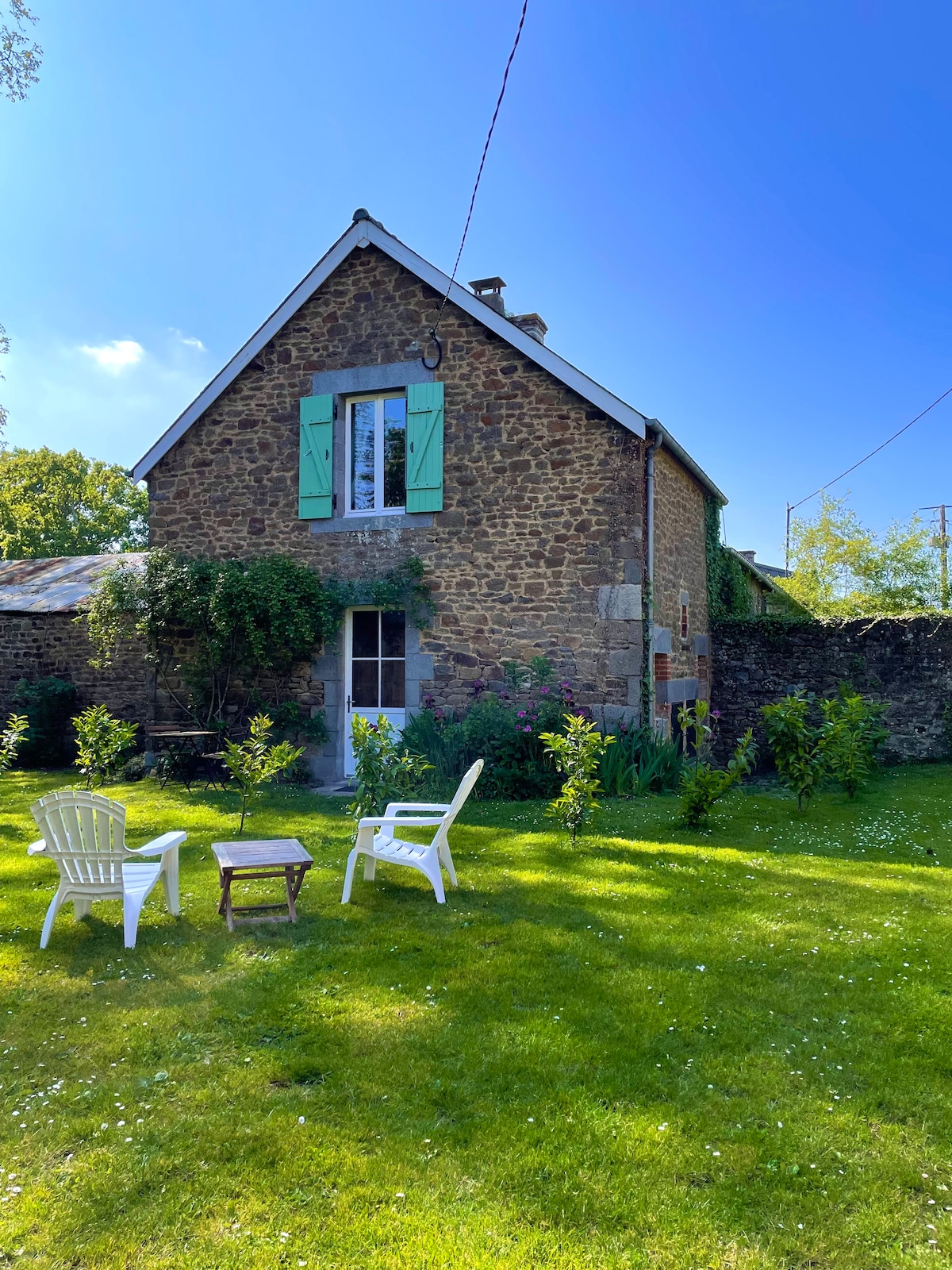 The exterior of the cottage is showcased, featuring stone walls and green shutters. A well-maintained garden is present, with a grassy area hosting three chairs and a small table. Sunlight brightens the scene, contributing to a peaceful ambiance.