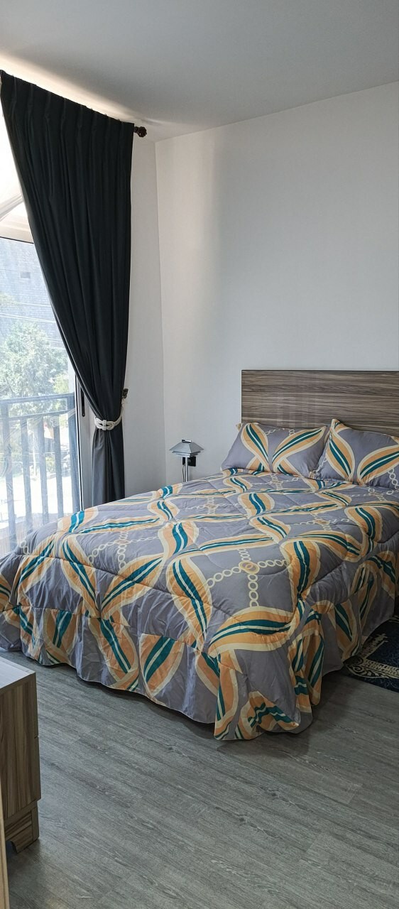 A well-arranged bedroom features a vibrant patterned bedspread covering a spacious bed. Natural light enters through a large window, draped with dark curtains. A bedside lamp provides illumination, while a wooden nightstand is positioned next to the bed, contributing to the room's functionality.