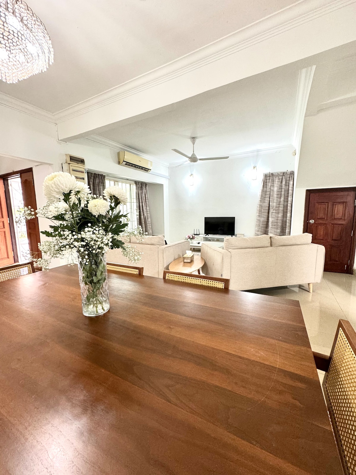A spacious living area is visible, featuring a comfortable sofa arrangement facing a wall-mounted television. Natural light fills the room through large windows adorned with light curtains. A dining table with a floral centerpiece sits in the foreground, offering an inviting ambiance.