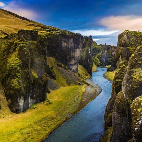 A scenic view of a winding river flows through a lush green canyon, bordered by towering cliffs covered in moss. The sky is filled with soft blue hues and wispy clouds, creating a serene atmosphere. Visitors can be seen in the distance along the ridge.