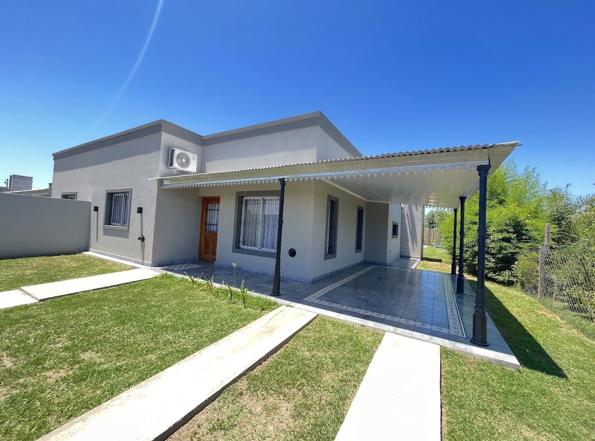 The exterior of the home is characterized by a light gray facade and a covered porch area. A manicured lawn is visible in the foreground, complemented by concrete pathways leading to the entrance. Large windows allow for ample natural light.
