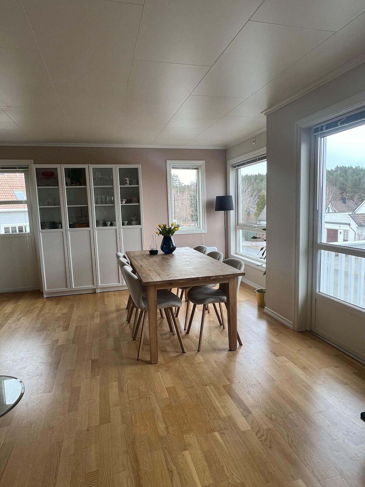 A bright dining area is shown, featuring a wooden table surrounded by light-colored chairs. Natural light streams through large windows, illuminating the pale walls and adding warmth. A modern lamp stands by a window, and a vase of flowers decorates the table.