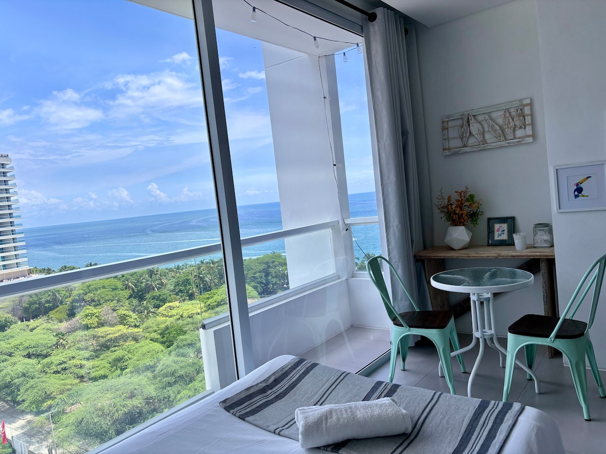 A bright and airy room features a spacious bed with soft linens. A small dining area with two chairs and a table is placed by the window, showcasing sweeping views of the sea and greenery outside. Natural light fills the space.