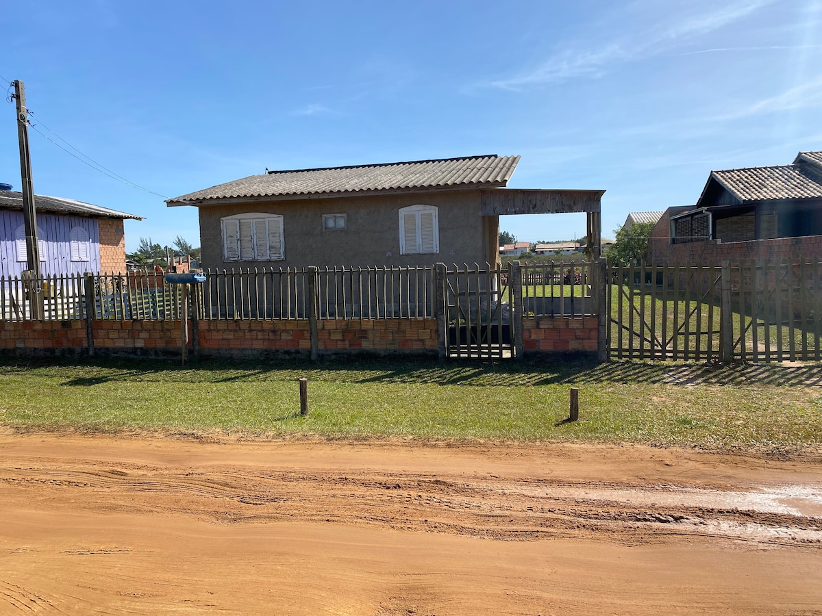 A small house is visible, featuring a covered porch with a wooden railing and white window shutters. A dirt path leads to the entrance, with a lawn area in front. Nearby structures are seen in the background, alongside a clear blue sky.