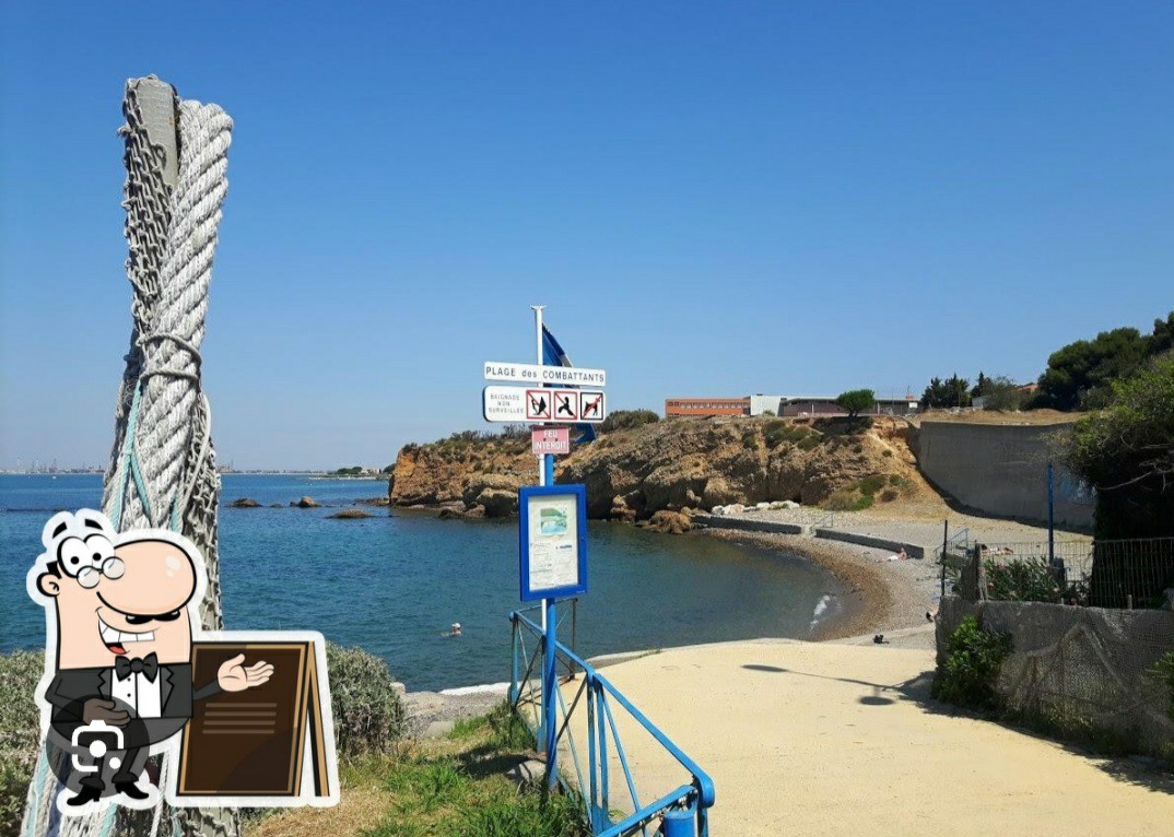 A serene coastal view is presented, featuring a sandy path leading to a pebbled beach. Clear blue skies stretch overhead, while signs indicate various regulations. The rocky shoreline and gentle waves contribute to the tranquil atmosphere of the area.