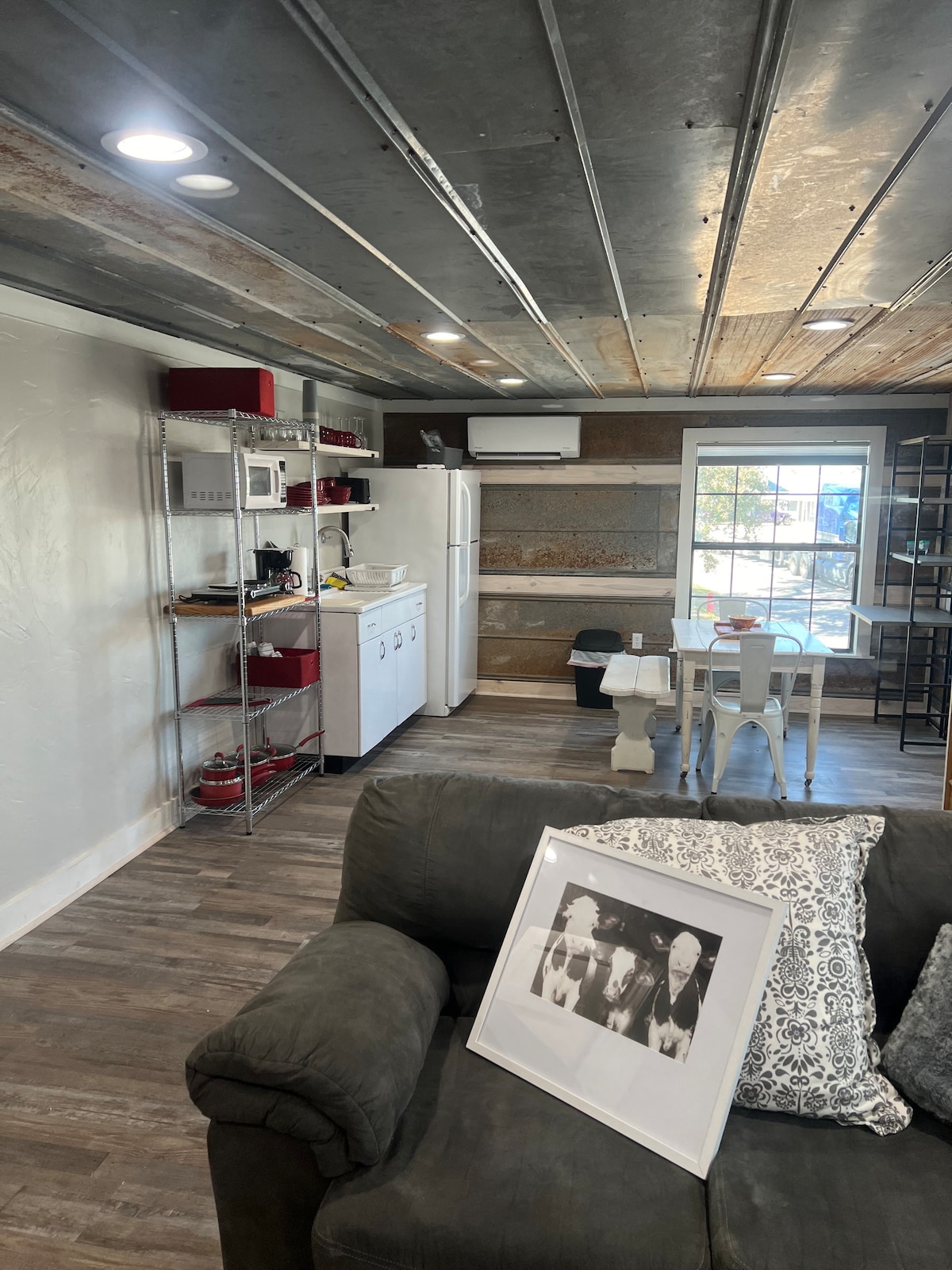 An open layout kitchen and living area is presented, featuring a spacious couch with decorative pillows. Metal shelving holds kitchen appliances and supplies, while a white dining table with chairs stands in the foreground. A window allows natural light to fill the space.